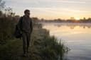 An angler walking a carp lake bank at first light, watching the water before setting up