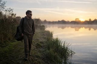 An angler walking a carp lake bank at first light, watching the water before setting up